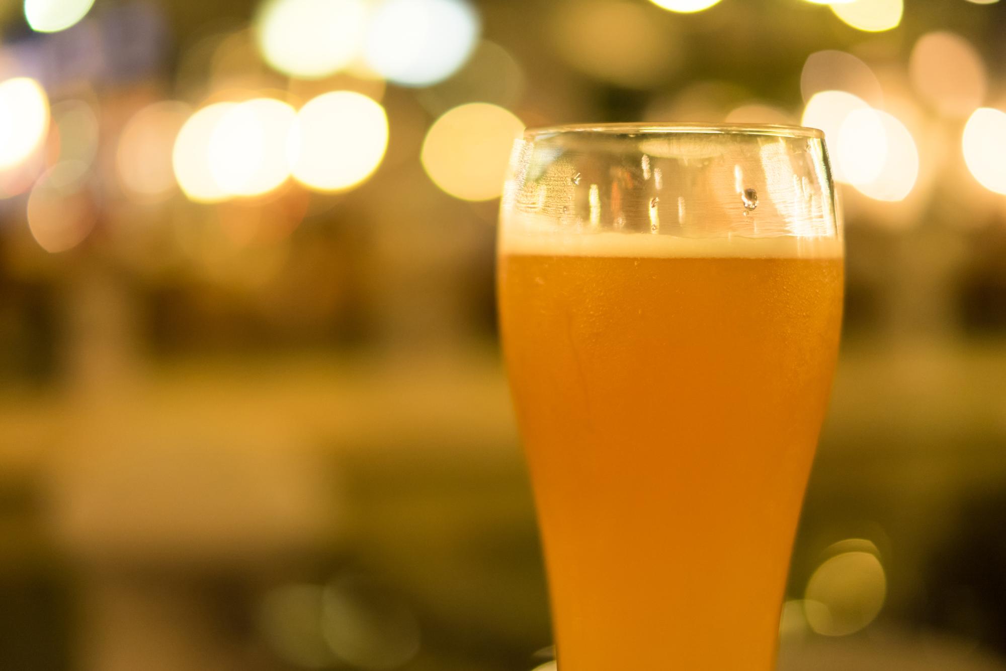 黄金色のクラフトビールが注がれたグラス。背景には温かみのある光のボケが見える、居心地の良いバーの雰囲気。